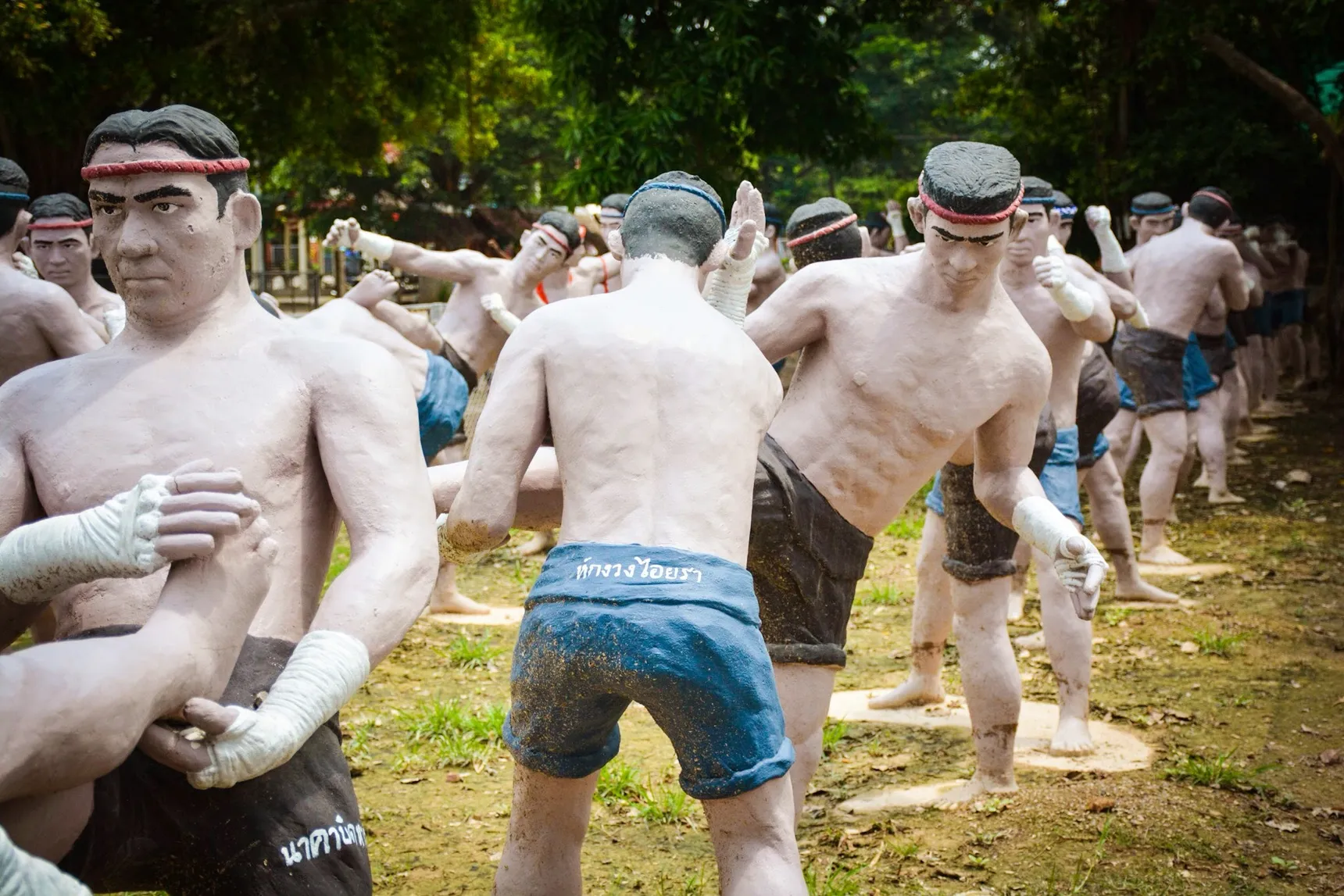 Life-size painted concrete statues of Muay Thai fighters at Bang Kung Camp in Samut Songkhram, Thailand, standing on grass under trees. Each is frozen mid-technique with rope-wrapped hands and a head band, and the name of the technique is painted in Thai script across the waistband of the shorts.